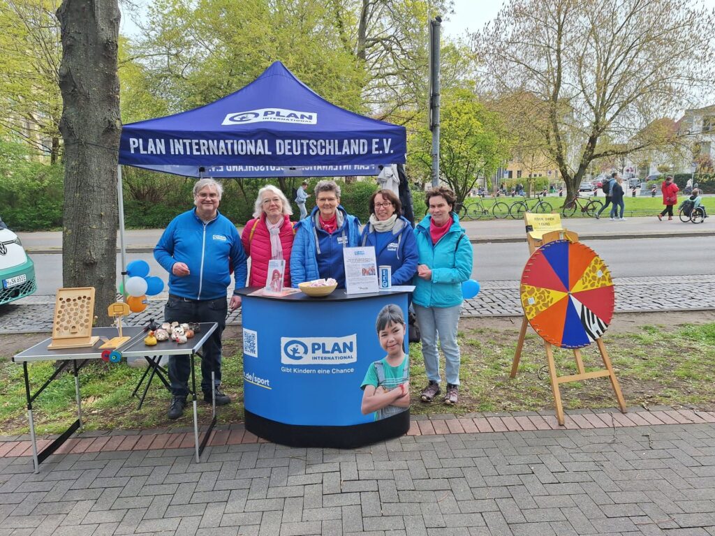 Five people stand at a Plan International Deutschland E.V. booth outdoors, with brochures, balloons, and a spinning wheel. The booth is set up on a sidewalk with trees and a street in the background.