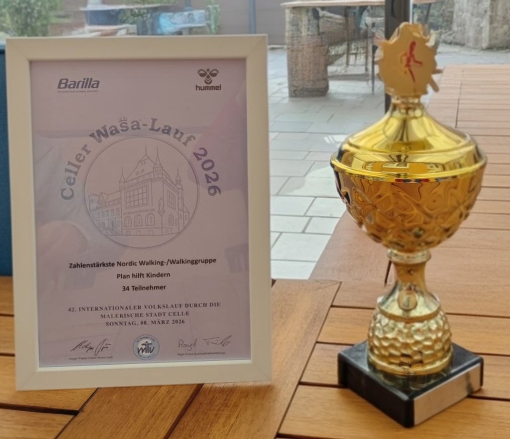 A framed certificate and a gold trophy with a running figure on top are displayed on a wooden table. The certificate is for the Celler Wasa-Lauf 2026 Nordic Walking group, awarded for participation.