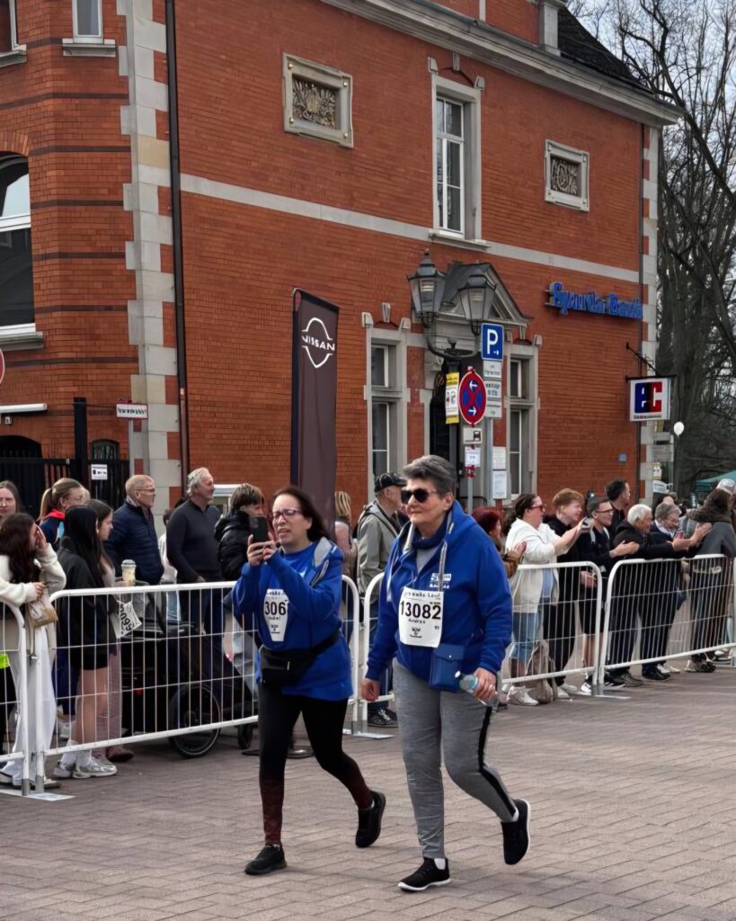 Two women wearing blue jackets and race bibs walk on a street during a public event, passing in front of a red brick building, while a crowd watches from behind metal barriers.