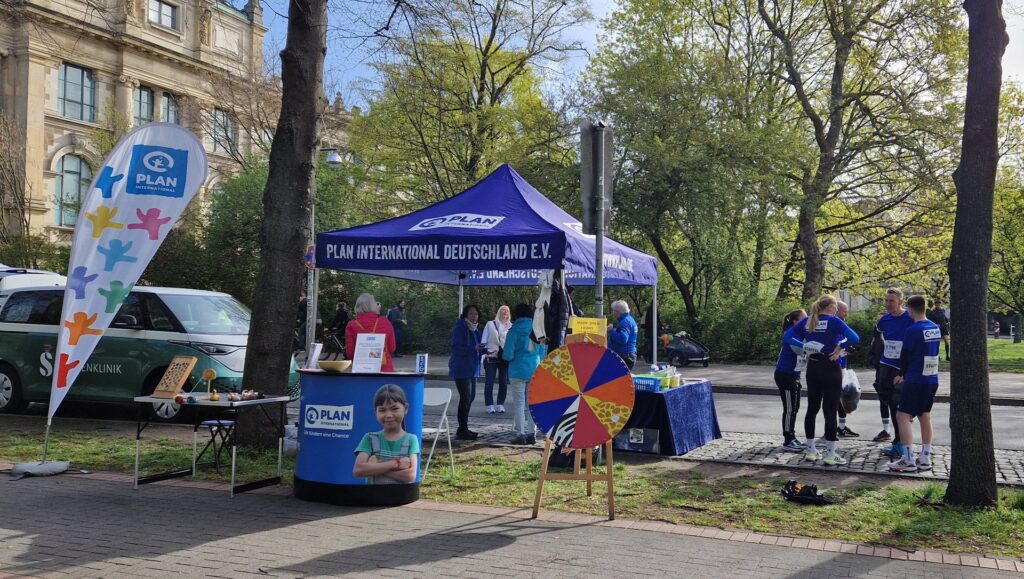 An outdoor event booth with a blue “Plan International Deutschland” tent, a display table, a colorful prize wheel, and several people standing and talking on a tree-lined street.