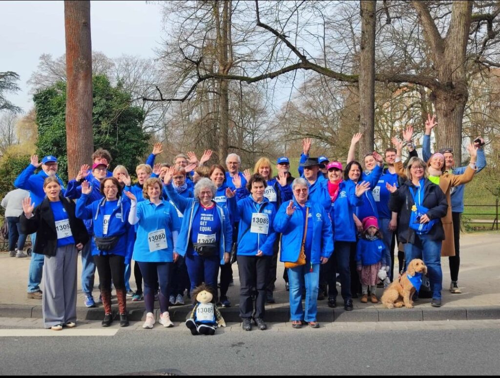 A large group of people wearing blue jackets and numbered race bibs stand on a sidewalk, smiling and waving at the camera. Some trees can be seen in the background, along with a small dog and a teddy bear in front.